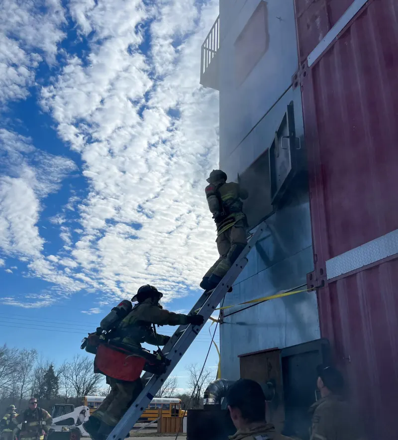Firefighter in full gear climbing a ground ladder against a blue sky